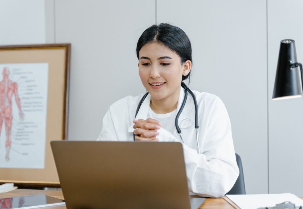 Nurse in a white coat and stethoscope smiling as she studies at her laptop