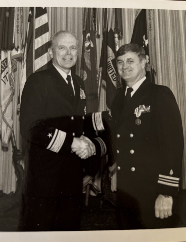 Black-and-white photograph of Capt. M. Ron Eslinger shaking hands with a Navy rear admiral at an awards ceremony, flags behind them